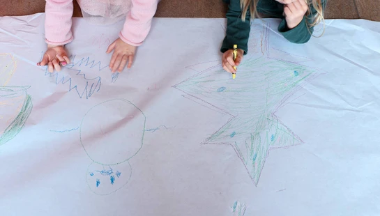 An overhead view of two little girls lying on the floor drawing with crayons on a large piece of white paper, one coloring in a star.